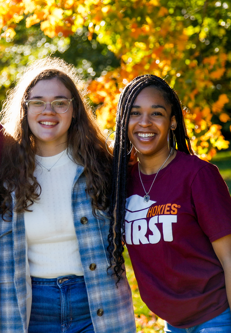 Two students stand side-by-side, arms around each other's back, with orange, red, and green foliage behind them.